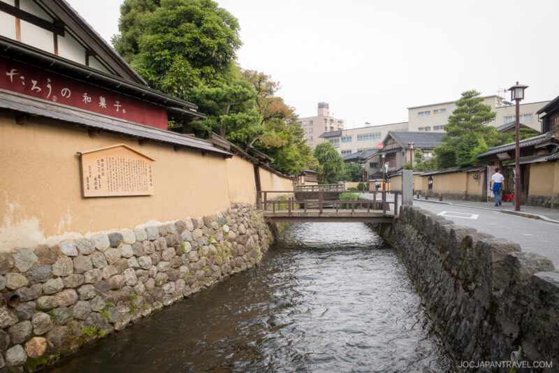 A narrow canal runs alongside traditional Japanese buildings in the Nagamachi Samurai District, with beige walls, stone foundations, and trees. A small wooden bridge crosses the canal as a few people walk on the quiet street nearby.