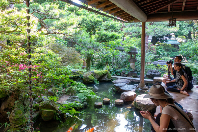 People sit on a wooden porch in the Nagamachi Samurai District, overlooking a serene Japanese garden with a pond, koi fish, lush greenery, and stone lanterns. One person takes photos while another looks at their phone.