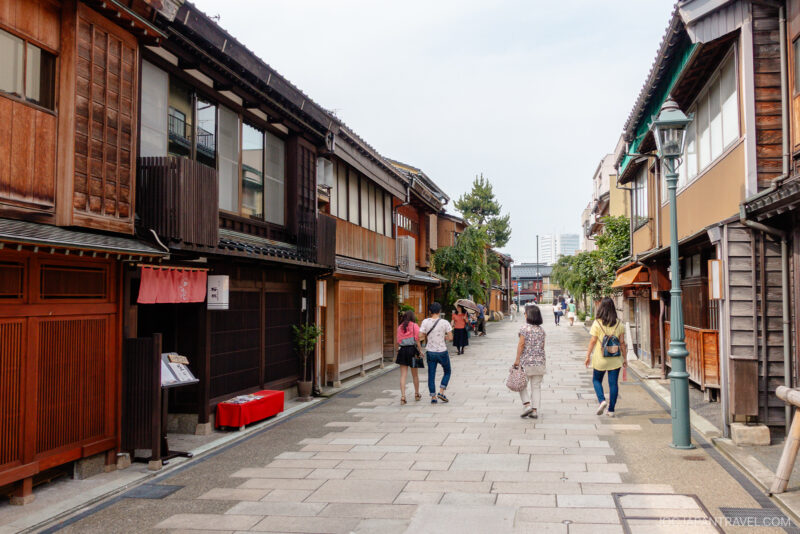 A stone-paved street lined with traditional wooden buildings in Japans Nishi Chaya District, with several people walking and enjoying the scenery under a bright sky.