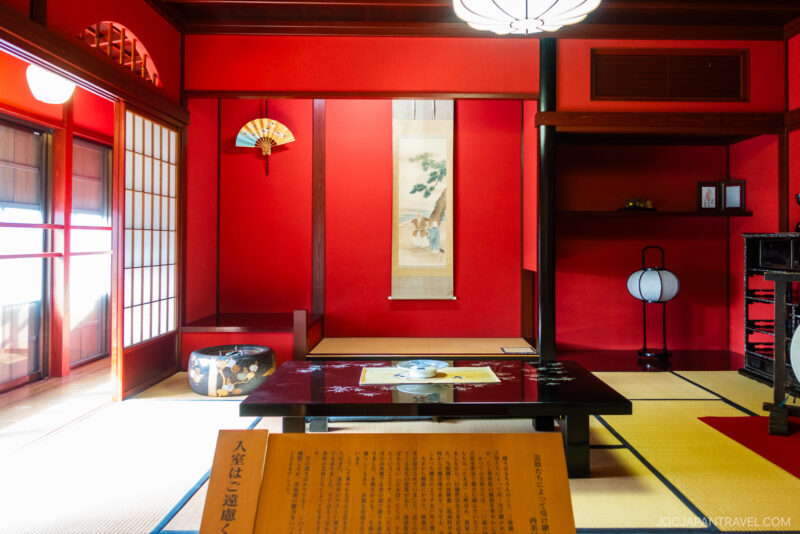 A traditional Japanese tatami room inspired by the Nishi Chaya District, with red walls, a low black table set for tea, hanging scroll, decorative fan, shoji screens, and soft natural light filtering in from the left.