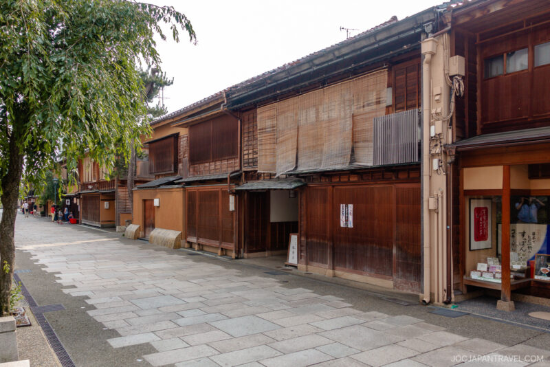 A quiet street in the Nishi Chaya District with traditional wooden buildings in Japan, featuring sliding doors, lattice windows, and bamboo blinds, with a tree and stone-paved sidewalk in the foreground.