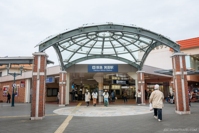 People walk near the entrance of Hankyu Minoh Station in Japan, which features a glass and metal canopy supported by brick columns. The station is a popular starting point for Osaka autumn foliage excursions. The station sign is visible above the entrance.