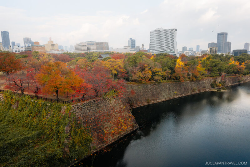 Cityscape with modern buildings in the background, Osaka autumn foliage of red and yellow leaves lining a stone wall along a river or moat under a partly cloudy sky.