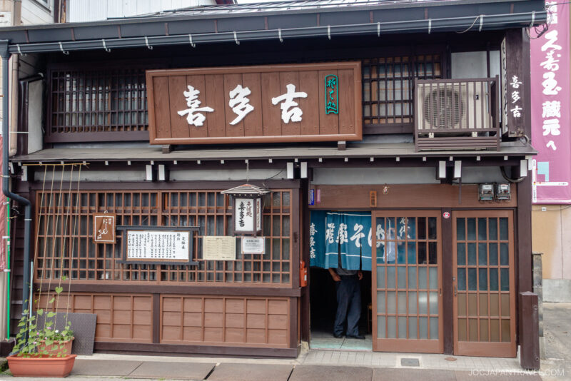 A traditional Japanese wooden storefront in Sanmachi Suji with sliding doors, lattice windows, and white kanji signage. Blue noren curtains hang at the entrance, partially revealing people inside. A potted plant sits by the door.