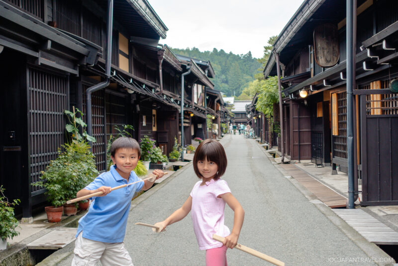 Two children stand on a quiet street in Sanmachi Suji, lined with traditional wooden buildings, holding wooden sticks and posing playfully. The scene is peaceful, with greenery and mountains visible in the background.