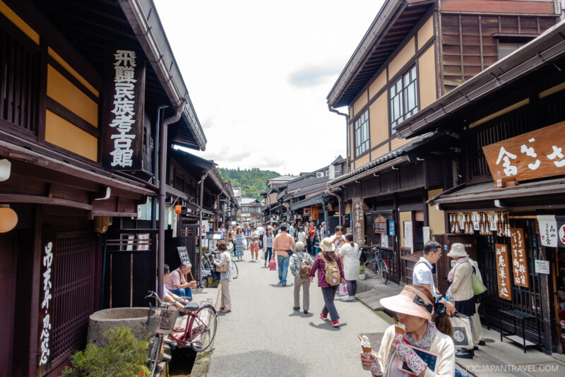 A lively street scene in Sanmachi Suji, a traditional Japanese town, with people walking and browsing shops housed in wooden buildings under a bright sky. Signs in Japanese hang from the buildings.