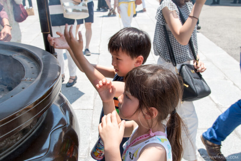 Two young children stand near a large incense burner outdoors, reaching toward the rising smoke with their hands. People are visible in the background on a sunny day.