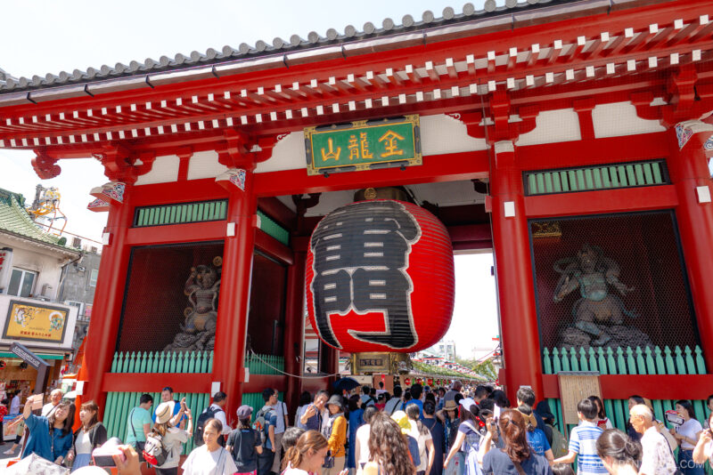 A large red lantern hangs at the entrance of Tokyo’s Senso-ji Temple, framed by bold red columns and a crowd of visitors below. Statues are visible in alcoves on either side of the gate.