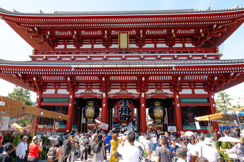 Large crowd gathers in front of the vibrant red and white Thunder Gate (Kaminarimon) at Senso-ji Temple in Tokyo, Japan, with large lanterns hanging beneath the ornate roof on a sunny day.