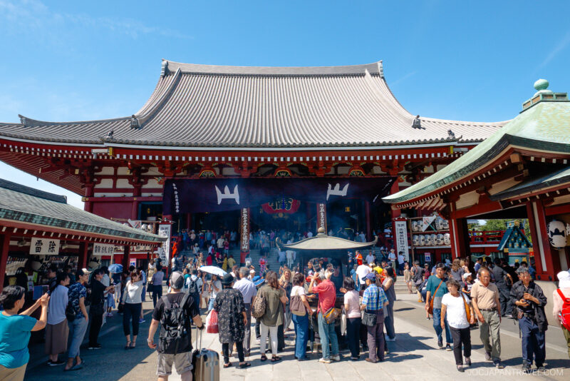 A large crowd gathers in front of the historic Senso-ji Temple in Tokyo, Japan, beneath ornate traditional roofs, with smoke rising from a central incense burner under a sunny sky.