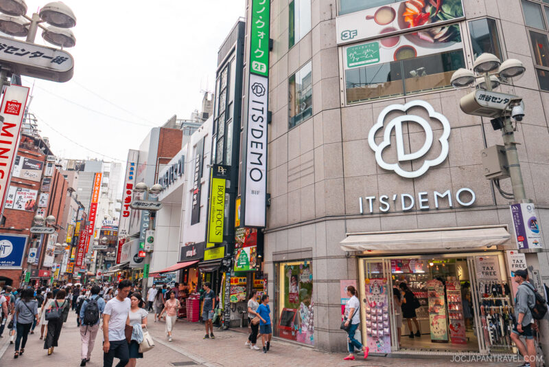 A busy street in Japan’s Shibuya Center Gai with people walking, colorful shop signs, and a large “IT’S DEMO” store on the corner. The lively atmosphere is filled with various shops and vibrant advertisements.