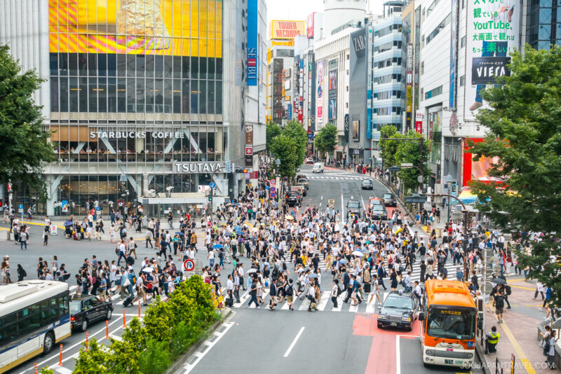 A busy crosswalk in Shibuya, Tokyo, with crowds of people crossing the street in multiple directions, surrounded by tall buildings, shops, bright signs, and vehicles. Experience the iconic energy of Shibuya’s bustling cityscape.