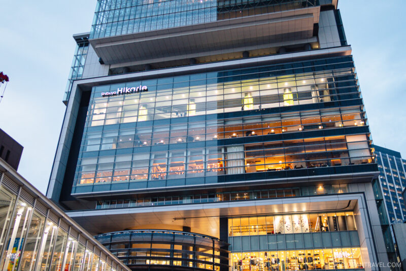 The modern multi-story Shibuya Hikarie features large glass windows, a brightly lit interior, and people visible inside. The building boasts a sleek, contemporary design in the heart of the city.