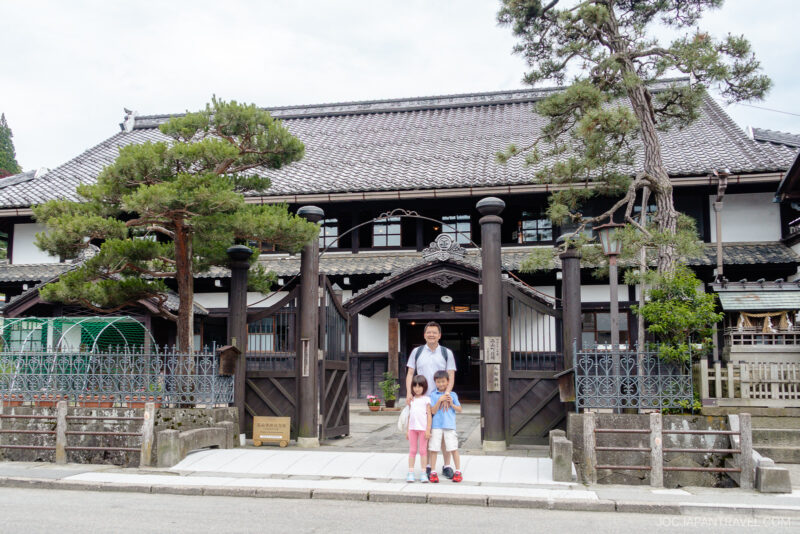 A woman and two children stand smiling in front of the Takayama City Archives Museum, a traditional Japanese wooden building with a tiled roof, surrounded by trees and a gated entrance on a cloudy day.