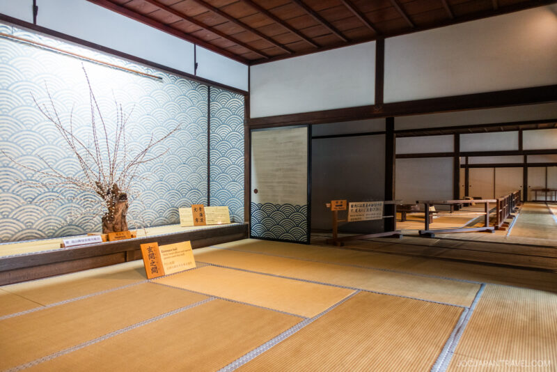 Traditional Japanese tatami room at Takayama Jinya with low partitions, sliding doors, a decorative wall, and a flower arrangement in a vase. Informational signs are placed on the floor, creating a calm and orderly atmosphere.