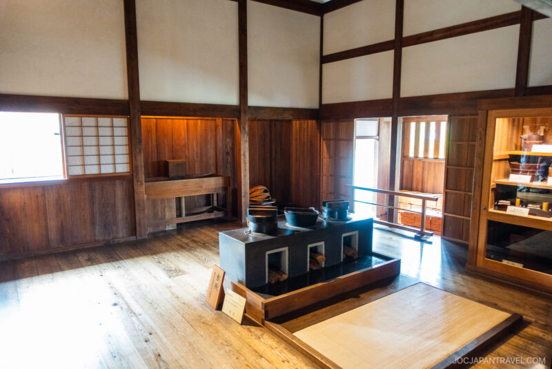 Traditional Japanese kitchen with wooden walls and floors, featuring a sunken hearth with three cooking pots—reminiscent of historic Takayama Jinya—shelves with utensils, and natural light streaming through a window.