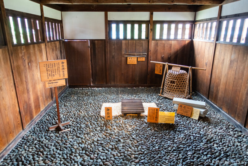 A traditional Japanese prison cell at Takayama Jinya with wooden walls, barred windows, a rock-covered floor, informational signs, and a bamboo cage in the corner.