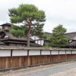 Traditional Japanese buildings with white walls and dark wooden frames stand behind a wooden fence at Takayama Jinya. Tall green pine trees rise above the structures, and a gravel path lines the foreground under a cloudy sky.