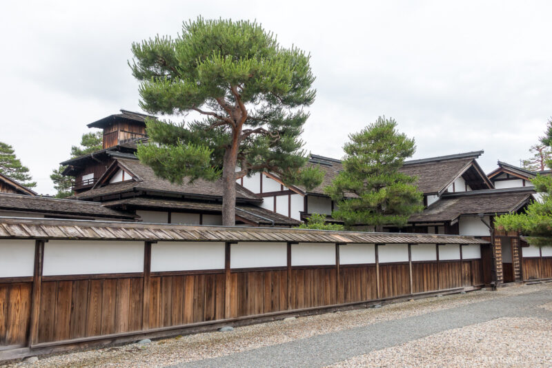 Traditional Japanese buildings with white walls and dark wooden frames stand behind a wooden fence at Takayama Jinya. Tall green pine trees rise above the structures, and a gravel path lines the foreground under a cloudy sky.
