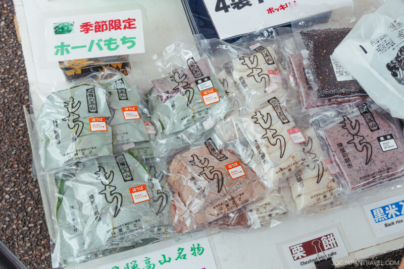 Individually packaged mochi in various flavors are displayed on a table for sale at the Takayama Jinya Asaichi outdoor market in Japan. Labels and colorful signs in Japanese provide information about the products.