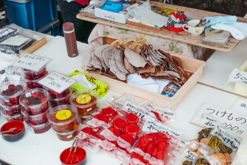 At Takayama Jinya Asaichi, a market stall displays Japanese pickled foods in plastic containers and bags, folded leaves, and handwritten price signs on a white tablecloth. Bowls, a cup, and small decorative items are also on the table.