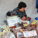 A vendor in a plaid shirt sits behind a market stall at Takayama Jinya Asaichi, displaying various packaged foods and handwritten price signs in Japanese. The goods are neatly arranged in plastic trays on a table.