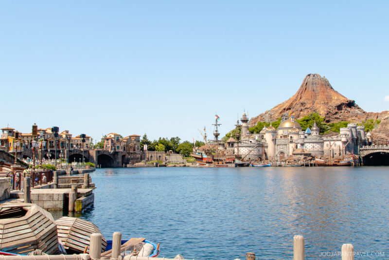 A wide lake with boats in the foreground, Mediterranean-style buildings on the left, and a volcanic mountain with a castle-like structure—iconic sights of Tokyo DisneySea—all under a clear blue sky.
