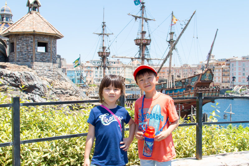 Two children stand smiling in front of a waterfront scene at Tokyo DisneySea, with a pirate ship and rustic buildings in the background on a sunny day. The girl wears a blue shirt, and the boy wears an orange shirt and red cap.