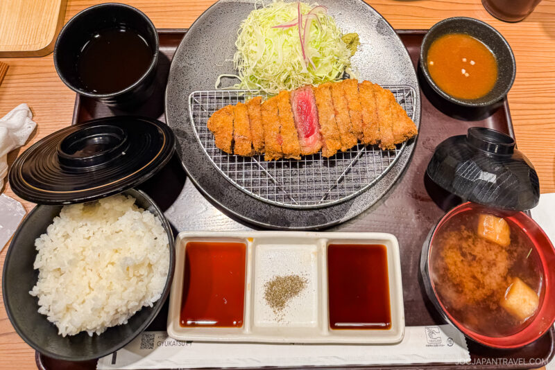 A Japanese meal set featuring sliced breaded pork cutlet, shredded cabbage, steamed rice, miso soup, dipping sauces, and a cup of tea—one of the must try foods in Tokyo—all served neatly on a tray.