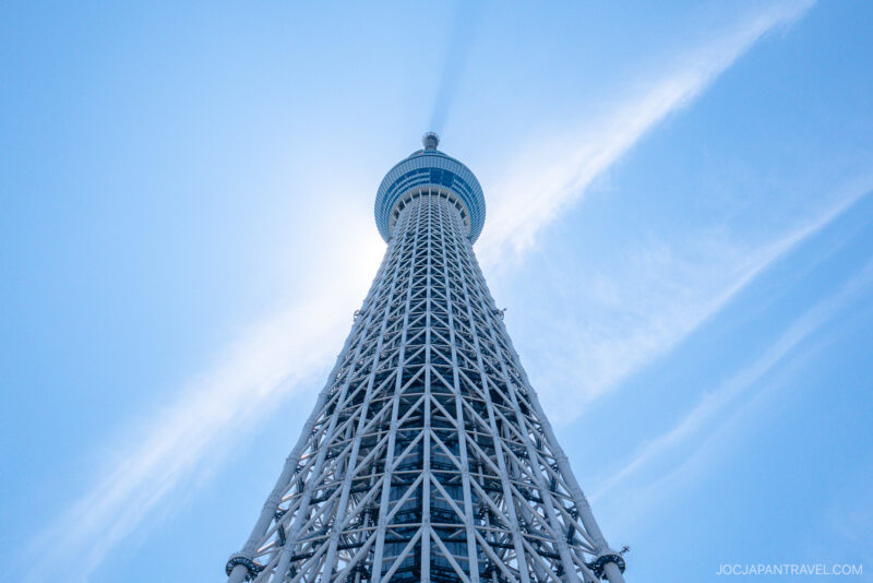 Tokyo Skytree tower photographed from below against a clear blue sky, with sunlight creating a halo effect around the top of Tokyo Skytree. Wispy clouds beautifully frame the towering structure.