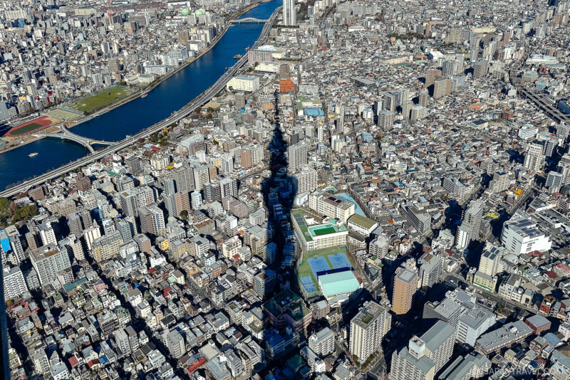 Aerial view of a densely packed cityscape with a river on the left; the tall shadow of Tokyo Skytree stretches across numerous buildings and streets in the sunlight.