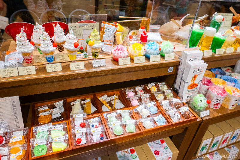 Display case with colorful, realistic plastic models of Japanese desserts like parfaits, ice cream, and sweets, alongside neatly arranged packaged treats in wooden trays below, in a bright, inviting Tokyo Skytree Town shop setting.