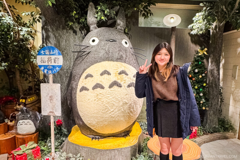 A young woman poses with a peace sign next to a large Totoro statue indoors at Tokyo Skytree Town, surrounded by plants and Christmas decorations. A smaller Totoro statue and a blue sign are visible to the left.