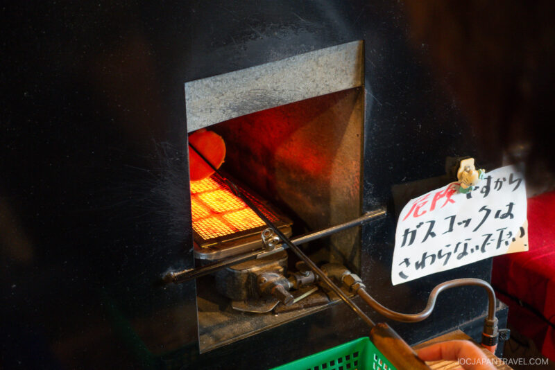 A person uses metal tongs to hold a round cracker over a hot grill inside an oven at Yume Kojo Hida. A handwritten sign in Japanese is attached to the oven’s side.