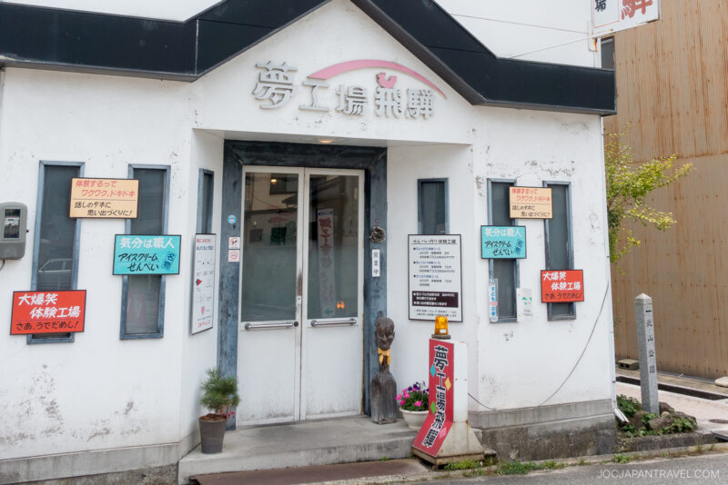 A small white building in Japan, Yume Kojo Hida, features multiple colorful Japanese signs around the entrance, a gray door with windows, a small plant, and a wooden statue near the door.