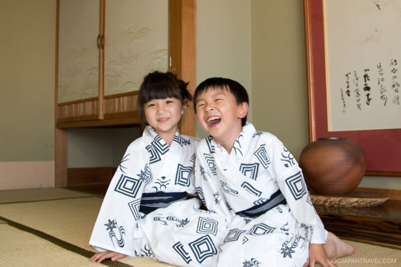 Two young children wearing matching white and black yukatas sit smiling and laughing on a tatami mat floor in a traditional Japanese room at Yunokawa Onsen, surrounded by wooden accents and a decorative scroll.