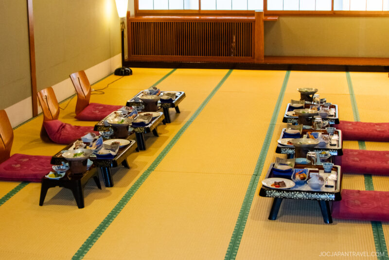 A traditional Japanese tatami room with low tables set for a meal, each with floor chairs and cushions. Various dishes are arranged on the tables, ready for dining. Warm lighting and shoji windows create a cozy atmosphere.