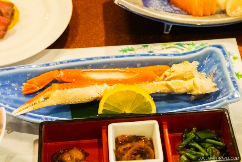 A blue dish with two chilled crab legs and a lemon slice, served alongside various small plates of vegetables and food at a Yunokawa Onsen ryokan.