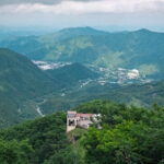 Akechidaira Plateau (明智平) – Nikko’s Most Breathtaking Scenic Route A scenic view of the lush green mountains of Nikko Akechidaira Plateau, featuring a small building and cable car station in the foreground, overlooking a valley with winding roads and distant villages under a cloudy sky.