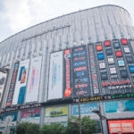 Large electronics store building near Akihabara Station in Tokyo, with multiple colorful signs and advertisements for gadgets, cameras, and phones. The vibrant, modern building is bustling, and most of the signage is in Japanese.