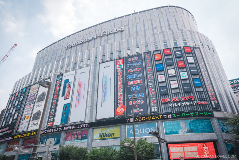 Large electronics store building near Akihabara Station in Tokyo, with multiple colorful signs and advertisements for gadgets, cameras, and phones. The vibrant, modern building is bustling, and most of the signage is in Japanese.