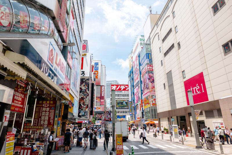 A busy city street near Akihabara Station in Japan, with tall buildings covered in colorful signs and advertisements, crowded with pedestrians under a bright, partly cloudy sky.