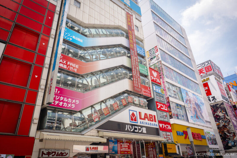 A busy street view near Akihabara Station in Tokyo, with tall, colorful buildings and the LABI electronics store displaying large Japanese signs, glass escalators visible, and a bright, clear sky above.