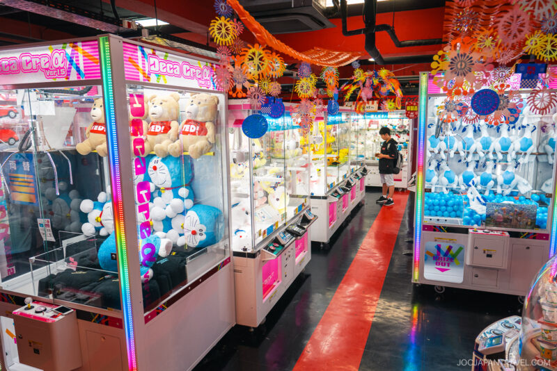 A brightly lit Akihabara Video Arcade filled with claw machines containing plush toys and balloons. Colorful decorations hang from the ceiling, and a person is seen playing one of the machines in the background.