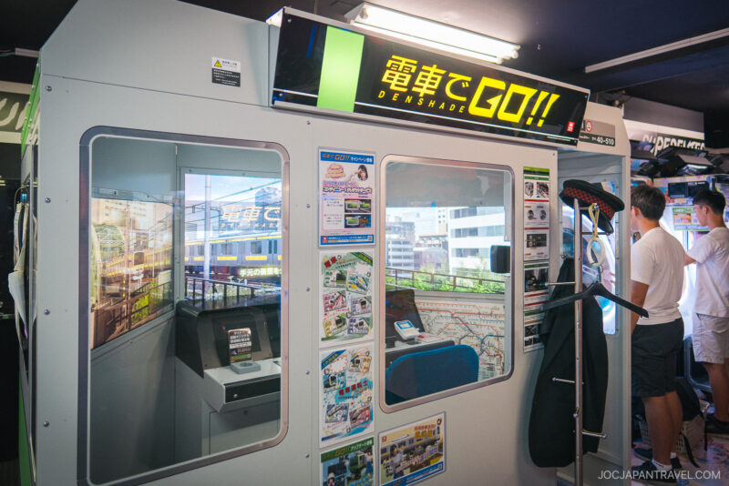 A Japanese arcade game booth labeled Densha de GO!! simulating train operation, with screens, controls, maps, and posters inside. Two people stand nearby in a brightly lit Akihabara Video Arcades center.