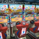 Four Mario Kart arcade racing game machines with red seats and steering wheels are lined up side by side in an Akihabara Video Arcades setting, each displaying colorful racing graphics on their screens.