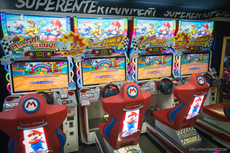 Four Mario Kart arcade racing game machines with red seats and steering wheels are lined up side by side in an Akihabara Video Arcades setting, each displaying colorful racing graphics on their screens.