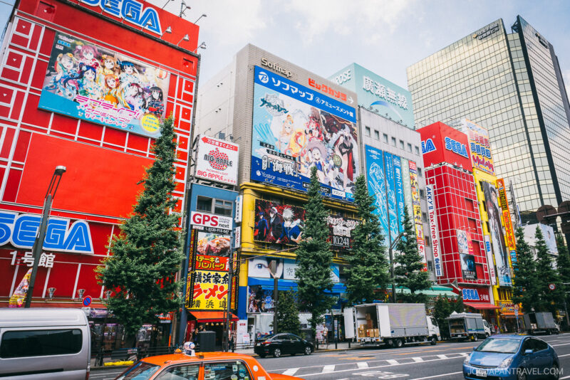 Colorful buildings in Akihabara, Tokyo, covered in anime and gaming ads—including large SEGA signs—line the street, where Akihabara video arcades buzz with activity among trees, cars, and pedestrians.