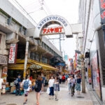 A busy pedestrian street in Ameyoko Market, Tokyo, with people walking past shops, restaurants, and colorful Japanese signs. The Ameyokocho entrance arch rises above the lively street scene.