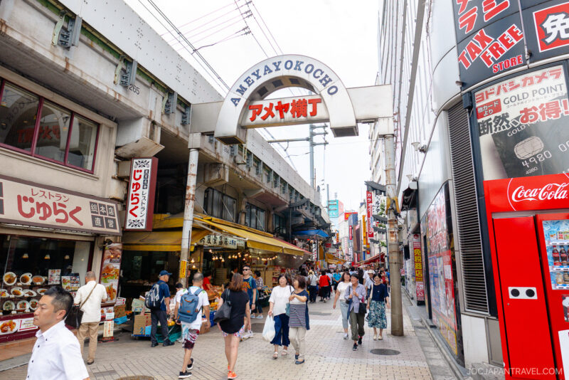 A busy pedestrian street in Ameyoko Market, Tokyo, with people walking past shops, restaurants, and colorful Japanese signs. The Ameyokocho entrance arch rises above the lively street scene.